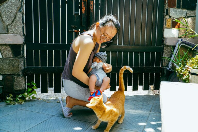 A woman in glasses holds a baby while interacting with an orange cat outside near a black gate.
